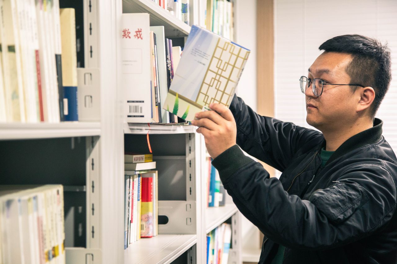 Asian man in glasses selecting a book from a library shelf, focused and thoughtful.